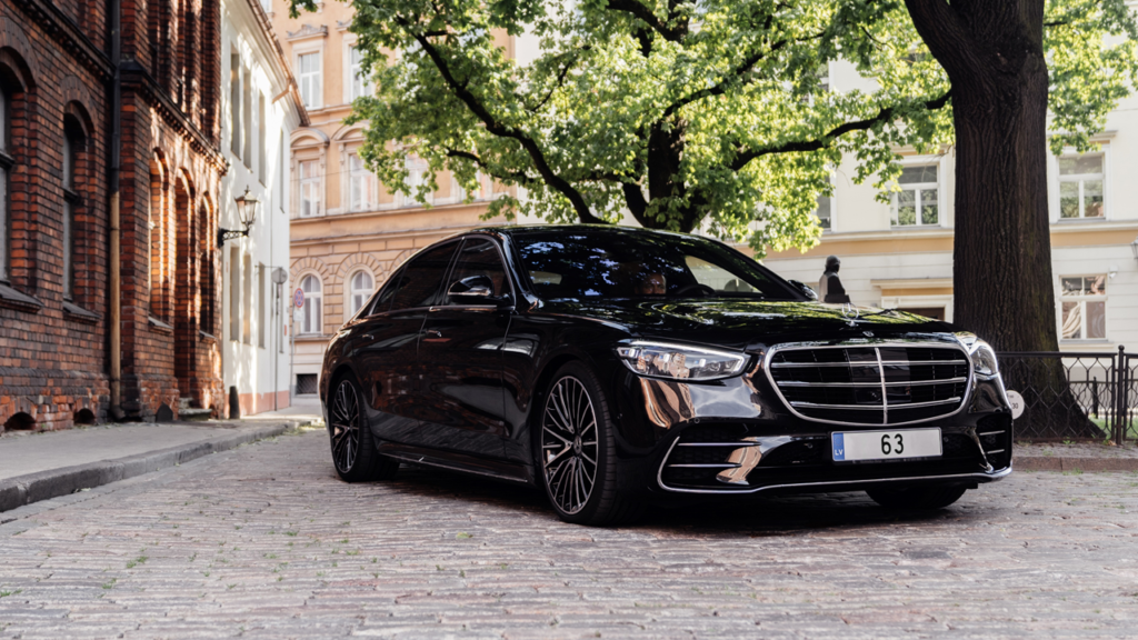 A sleek black Mercedes-Benz executive sedan parked on a cobblestone street alongside classic European-style brick architecture and lush green trees.