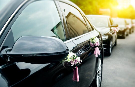 Row of black luxury sedan vehicles adorned with white and pink flower bouquets on door handles for a wedding procession.
