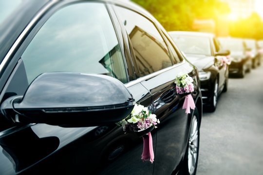 Row of black luxury sedan vehicles adorned with white and pink flower bouquets on door handles for a wedding procession.