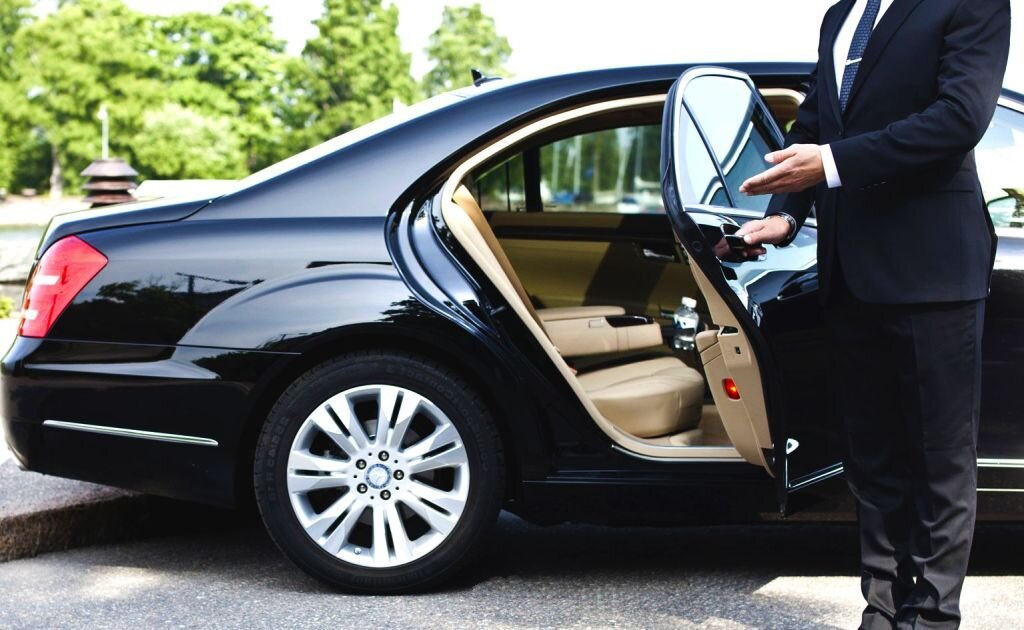 A professional chauffeur in a formal suit opening the rear door of a black luxury Mercedes-Benz sedan with tan leather interior.