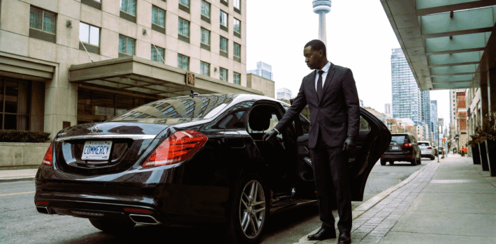A professional chauffeur wearing a suit opening the door of a black luxury Mercedes-Benz sedan parked on a Toronto street with the CN Tower in the background.