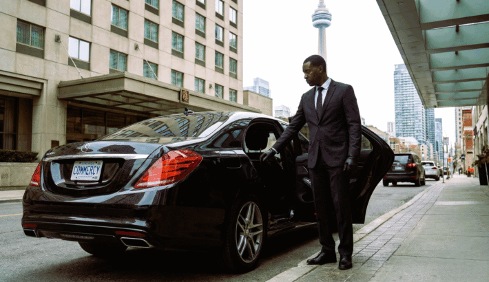 A professional chauffeur wearing a suit opening the door of a black luxury Mercedes-Benz sedan parked on a Toronto street with the CN Tower in the background.