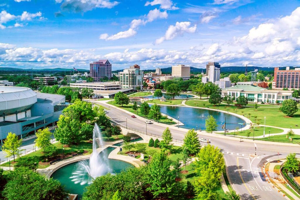An elevated, wide-angle shot of a lush green city park in Huntsville featuring a large fountain, walking paths, and ponds, with mid-rise office buildings and a clear blue sky in the background.
