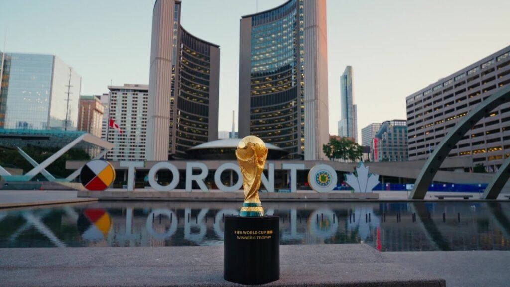 A high-angle, wide shot of Nathan Phillips Square in Toronto featuring the solid gold FIFA World Cup Trophy on a plinth in the foreground, with the curved towers of Toronto City Hall and the colorful "TORONTO" sign in the background.