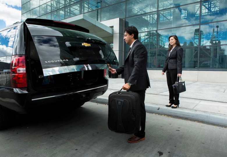 Executive-Corporate-Limo-Service-Canada Two corporate professionals in business suits loading luggage into the back of a black luxury Chevrolet Suburban SUV in front of a modern glass office building.