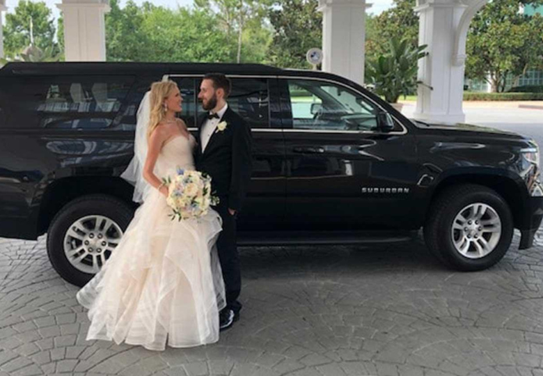 Luxury-Wedding-Limo-Service-Canada A bride in a white wedding dress and a groom in a black tuxedo standing in front of a black luxury SUV limousine under a white porte-cochère.