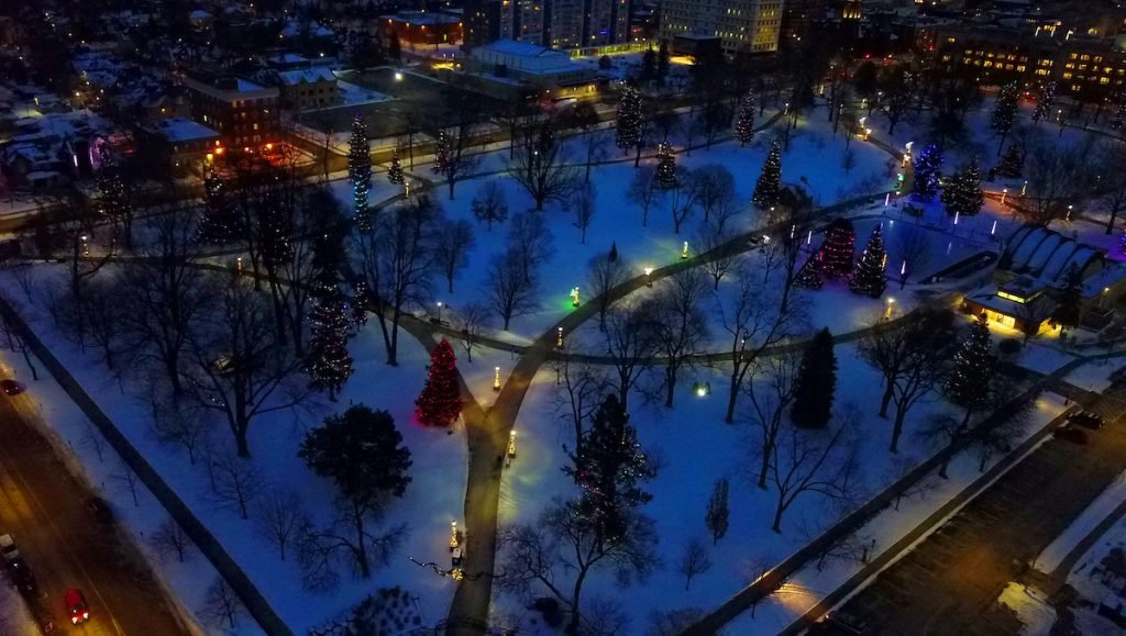 An aerial evening view of Victoria Park in London, Ontario, covered in snow. The park's winding pathways are illuminated by warm street lamps, and numerous evergreen trees are decorated with vibrant, colorful Christmas lights. The surrounding city buildings and streets are visible in the background under a dark twilight sky.