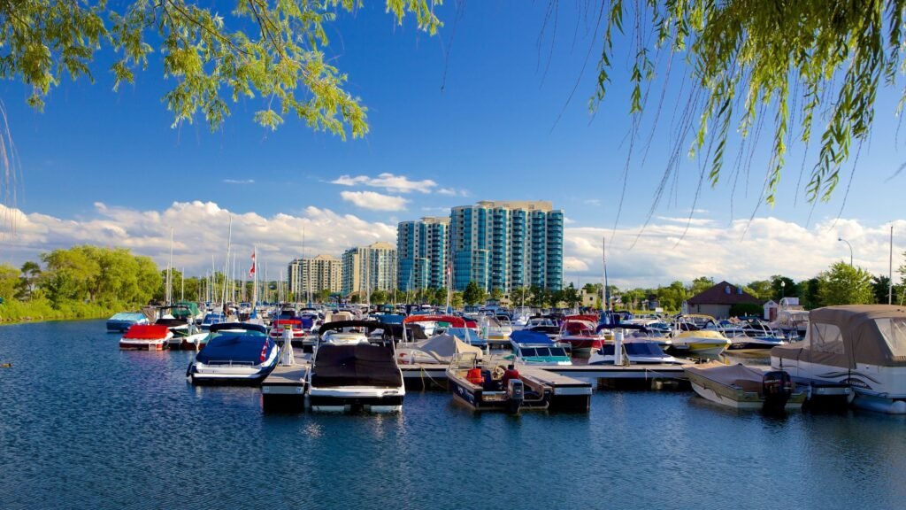 A vibrant sunny day at a marina in Orillia, Ontario, featuring numerous motorboats docked in blue water with modern high-rise residential buildings and lush green trees in the background under a clear blue sky.