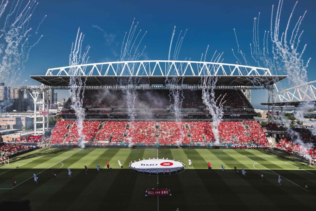 A wide-angle view of BMO Field in Toronto during a celebratory event with fireworks and smoke rising behind the stands. The stadium, set to host the FIFA World Cup 2026, features red seating filled with fans and a large BMO logo on the pitch.