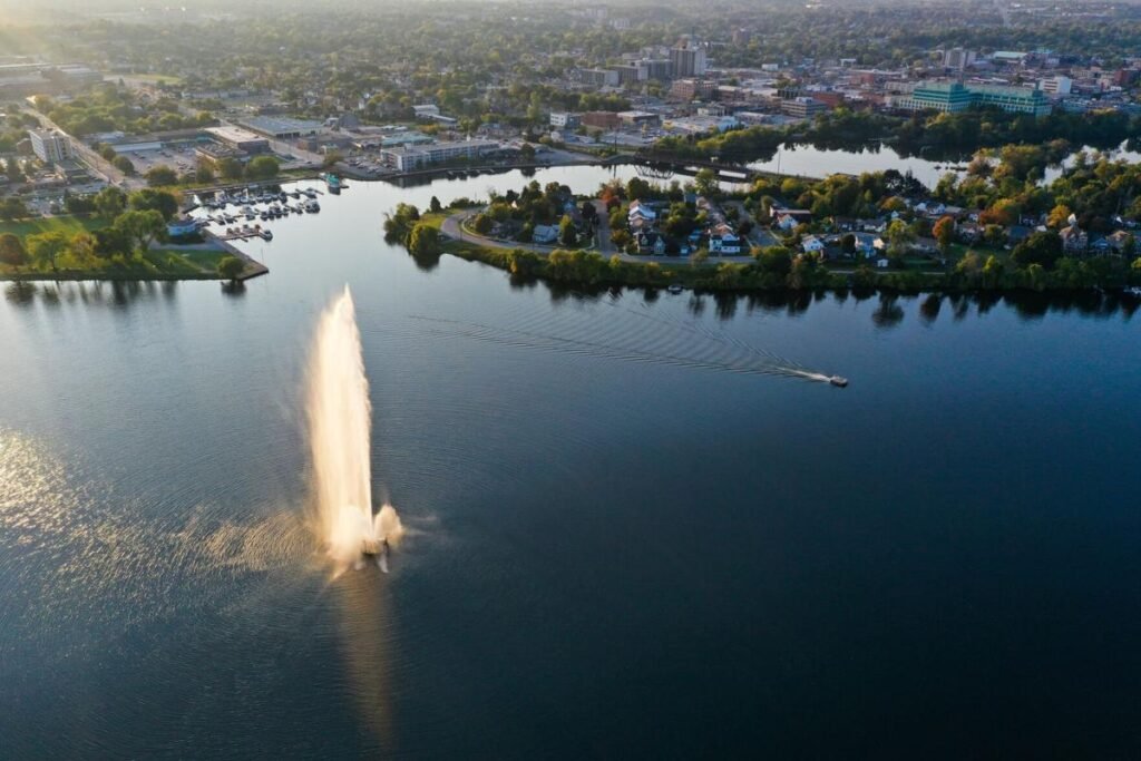 An aerial view of a large water fountain spraying high into the air on Little Lake in Peterborough, with the city skyline and marina in the background.
