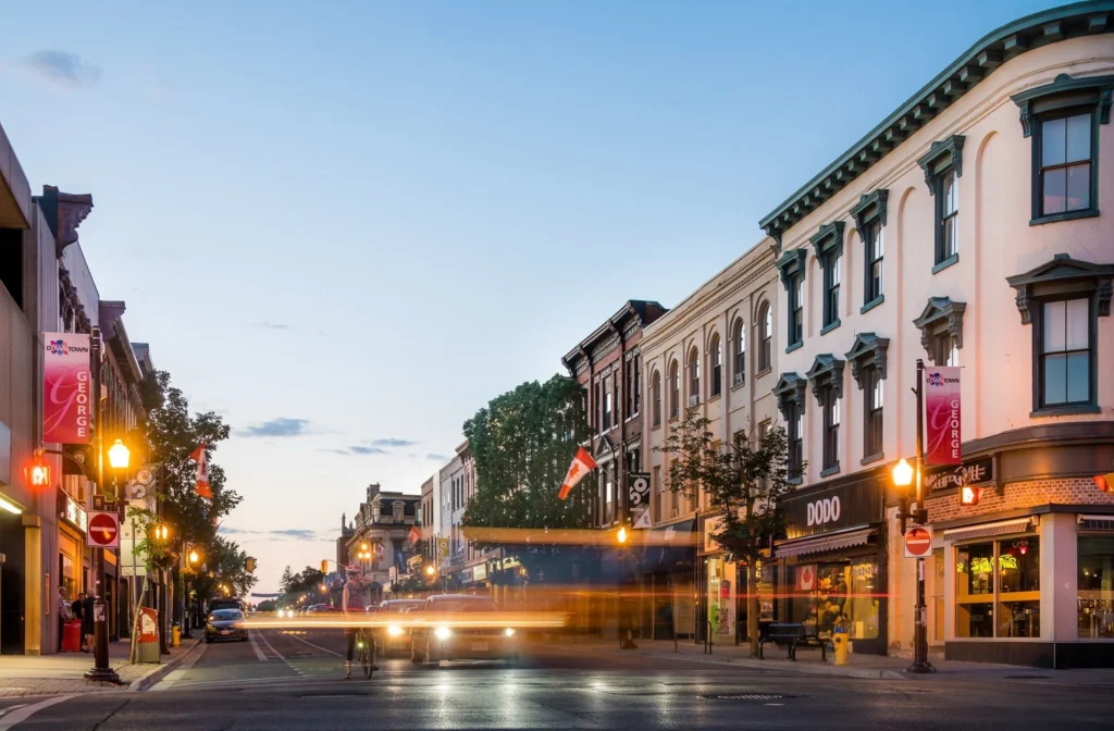 A street-level view of George Street in downtown Peterborough during dusk, featuring historic brick buildings, storefronts, street lamps, and light trails from passing cars.