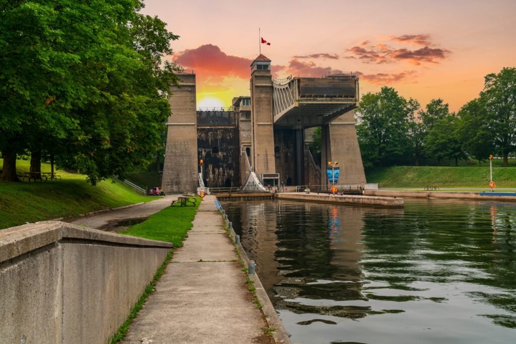 A large concrete hydraulic lift lock structure standing over a calm waterway in Peterborough, Ontario, framed by green trees under a vibrant warm sunset sky.